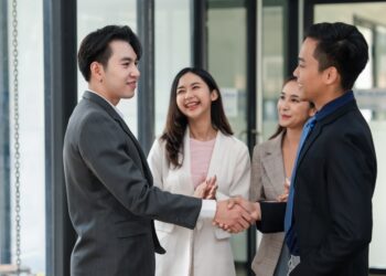 Two,Businessmen,Shaking,Hands,With,Colleagues,Applauding,In,Office,Environment.